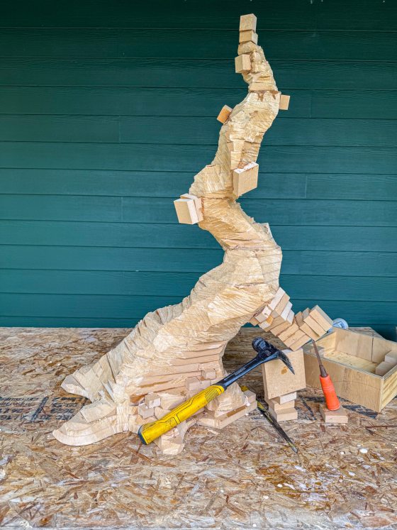 A rough-carved MDF model of a bonsai tree stands on a workshop table. A hammer and other carving tools lie nearby among wood shavings, showing the transition from square boards to a faceted organic shape.