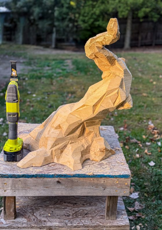 A carved wooden model of a faceted bonsai trunk sits on a wooden base. A yellow and black power tool stands upright next to the sculpture on a workshop table.