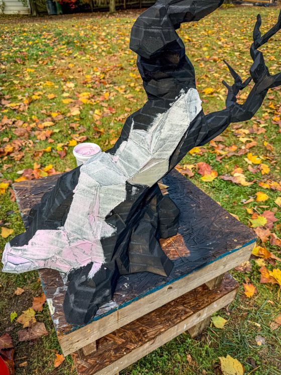 A large, black, geometric bonsai tree sculpture sits on a wooden platform outdoors. Sections of the faceted surface are covered in white and pink filler material as part of the smoothing process.