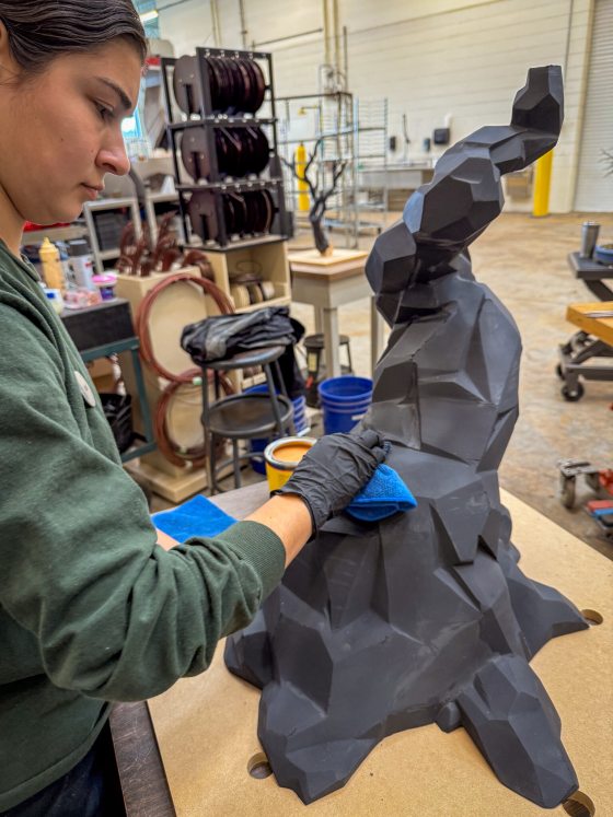 An artist wearing black gloves and a green shirt wipes down a large, black, faceted bonsai tree sculpture with a blue cloth in a workshop setting.