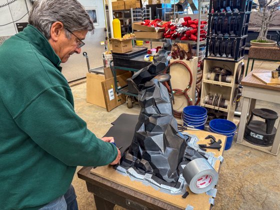 A man in a green fleece uses scissors to trim black foam around the base of a dark grey, faceted sculpture model on a workbench.