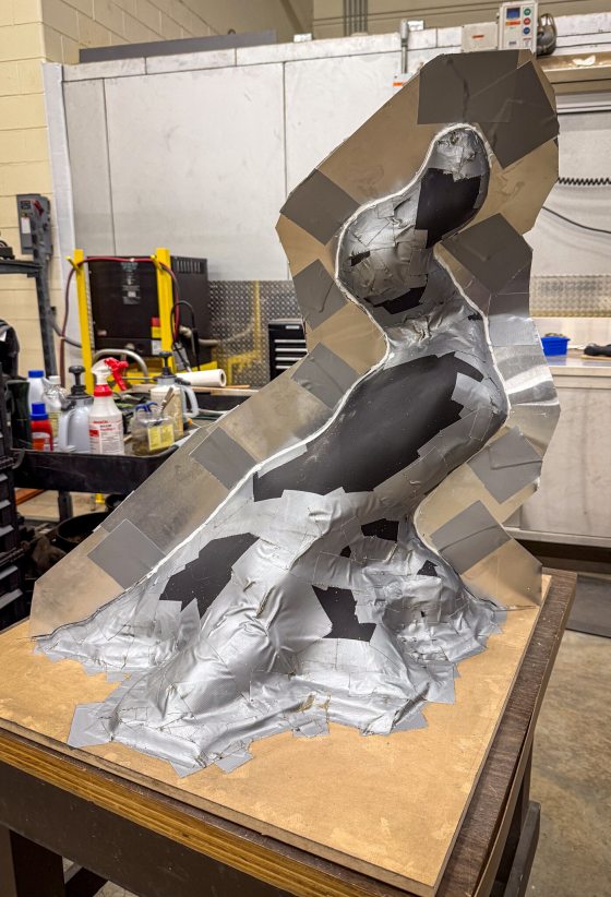 A black, angular sculpture model is prepared for mold-making, partially covered in silver duct tape and foil, standing on a wooden base in a workshop.