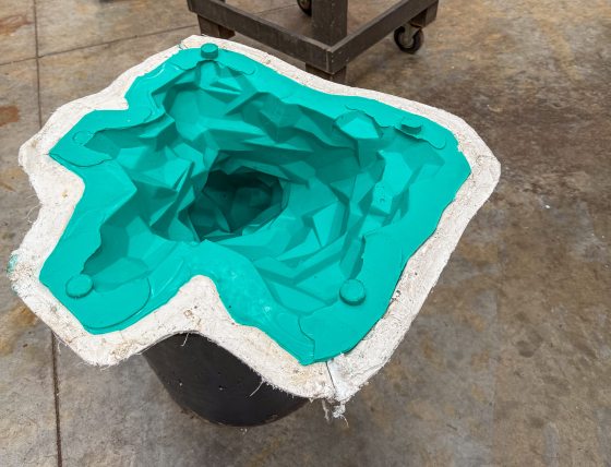 An open mold shows a bright teal silicone interior with a complex, faceted geometric cavity, supported by a white plaster outer shell.