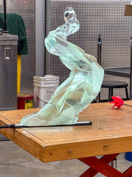 A translucent, light green faceted resin sculpture resembling a stylized bonsai trunk rests on a wooden workbench, illuminated from below by a black LED strip to highlight its crystalline structure.