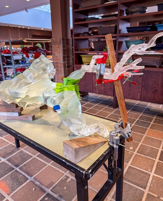 A large, multi-part translucent resin sculpture is held together with clamps and wooden supports on a workbench. Green and blue tape mark connection points on the faceted, crystalline branches during assembly.