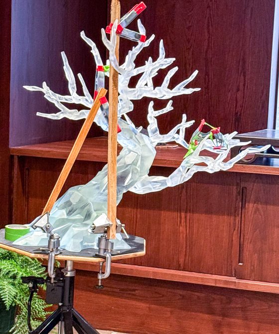 A completed, translucent faceted resin bonsai tree sculpture is secured to a rotating stand with clamps and wooden braces for final adjustments and finishing.
