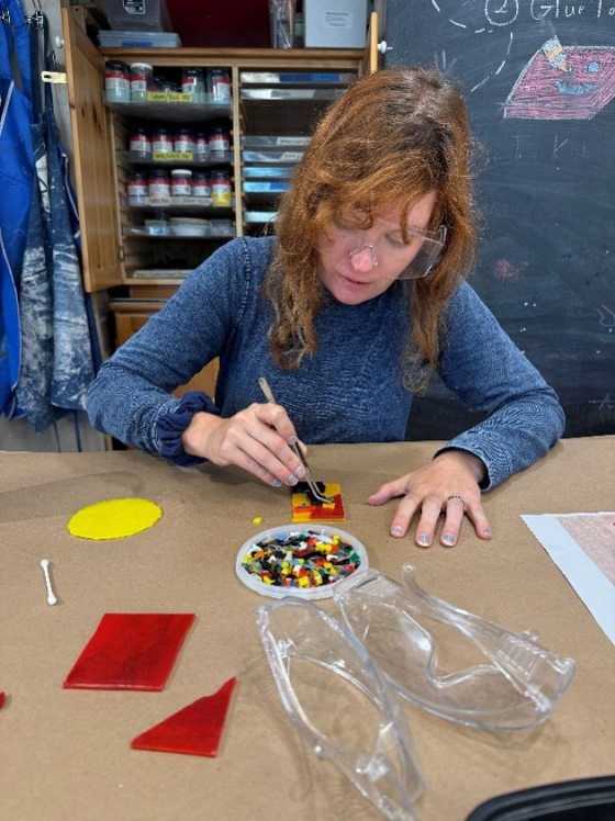 A woman with red hair and safety glasses is seated at a table covered in craft paper, working on a mosaic or fused glass project. She is using tweezers to place small, colorful glass pieces onto a base, with a dish of colorful pieces, a yellow circle, and large clear plastic molds nearby.