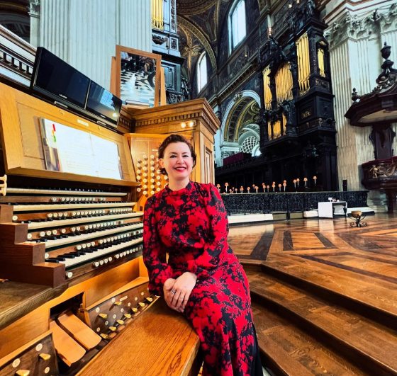 A person in a red and black dress half seated and leaning against an organ. 