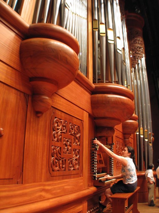 A person seated at a large organ, with silver pipes towering towards the ceiling. 