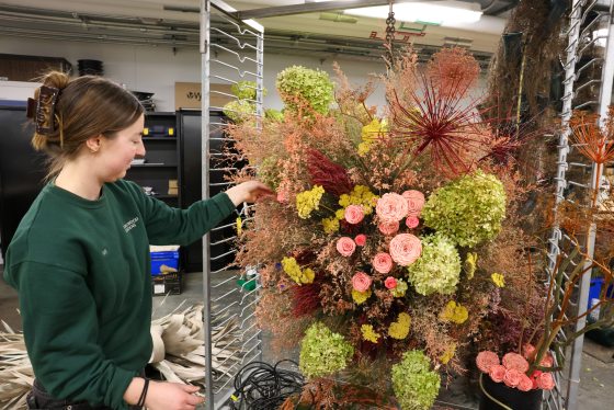 A staff member in a grey t-shirt focuses on assembling a lush arrangement of pink, white, and orange flowers on a work table.