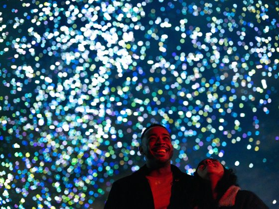 A close-up, low-angle shot of a man and woman looking upward with joy and wonder. The background is a dense, vibrant field of out-of-focus white, blue, and green light orbs that resemble a celestial event or a swarm of bioluminescent particles. The man’s face is illuminated by a warm red light from below.