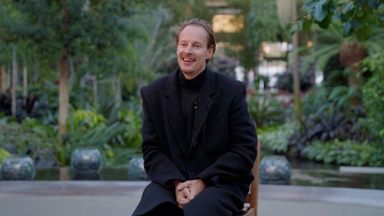 A person seated in a chair, in an all black suit, smiling and looking off into the left side of the photo.