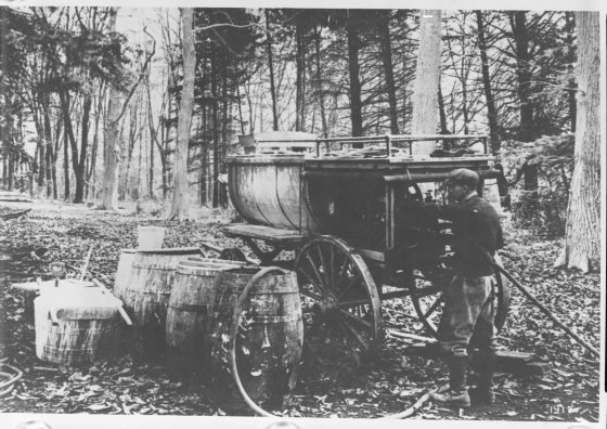 A black-and-white historical photograph from 1912 showing a man operating a large, horse-drawn wooden tank sprayer in a wooded area. Several wooden barrels and buckets are scattered on the leaf-covered ground nearby.
