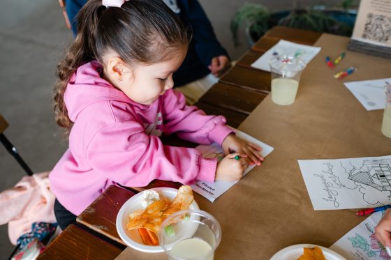 A young girl in a bright pink hoodie sits at a craft table, intently coloring a "Pileated Woodpecker" activity sheet with a green crayon.