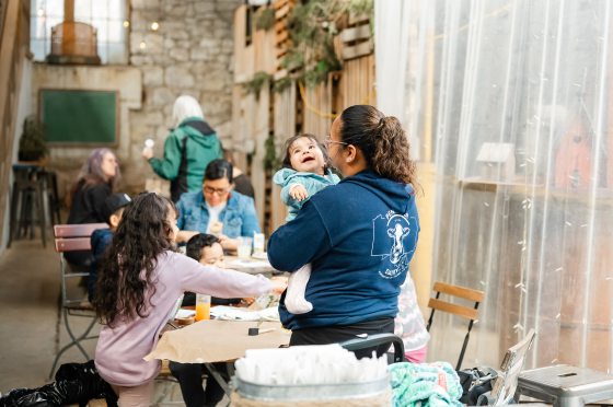 A wide shot of the community space showing a woman holding a smiling baby in the foreground, while families in the background engage in various crafting activities at long tables.