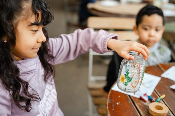 A young girl in a lavender sweatshirt holds up a clear glass jar she has decorated with delicate pressed flowers and green ferns.