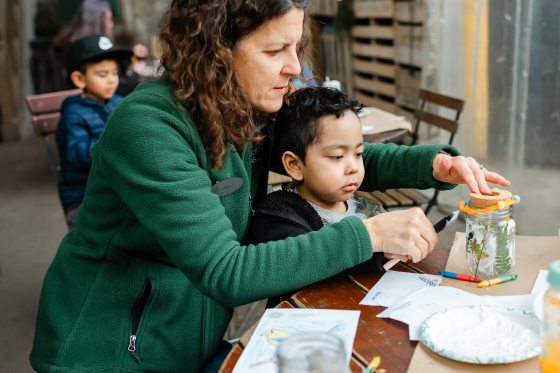An instructor in a green fleece leans in to help a young boy secure a cork lid and colorful beaded wire to the top of his finished decorative jar.