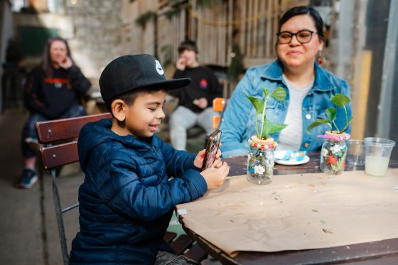 A young boy in a blue puffer jacket and black Nike hat sits at a workshop table, focused on taking a photo of a decorated glass jar with a smartphone.