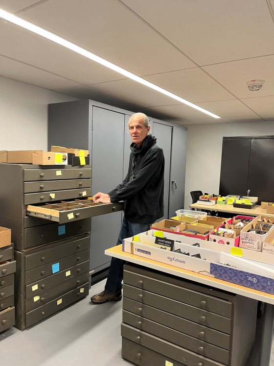 A man in a black hoodie stands in a storage room, pulling out a drawer from a large, olive-green industrial filing cabinet. The drawer is divided into small compartments filled with black plant labels. Various other boxes of labels, many with yellow adhesive notes for organization, are stacked on a nearby table and another filing cabinet.