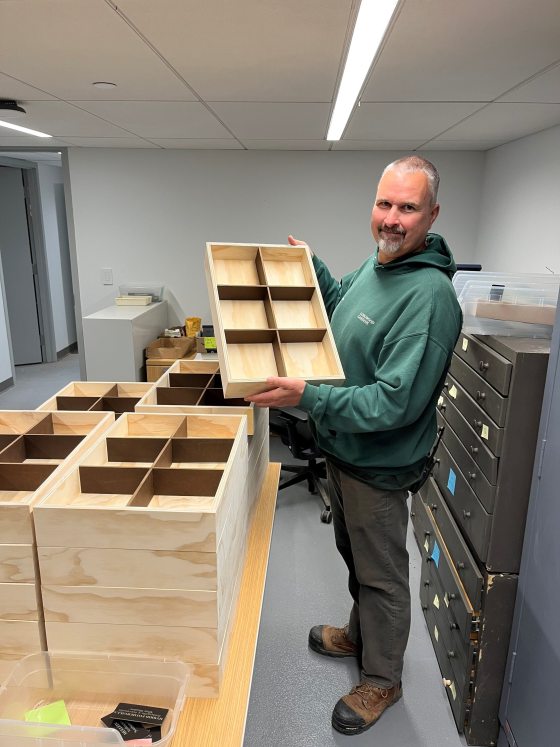 A man in a green Longwood Gardens sweatshirt holds up a handmade, light-colored wooden storage tray with six divided compartments. He is standing in a room with stacks of similar wooden trays and several large, metal filing cabinets used for storing plant identification materials.