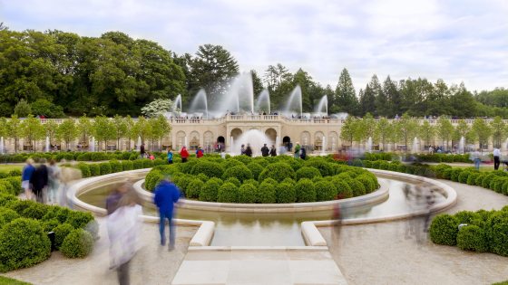 Visitors stroll through a fountain garden lush with green boxwoods and fountain jets shooting in the air