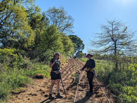 Two people stand on a dirt incline that has been prepared for planting with rows of small holes. One holds a long-handled tool, and the other holds a power drill. They are dressed in work clothes and hats under a bright, clear blue sunlit sky. A stone building is visible in the distance.