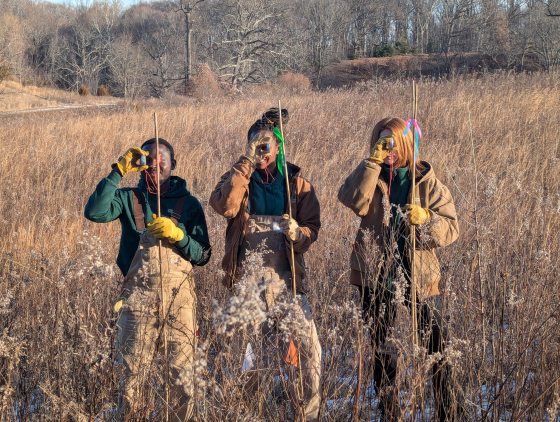 Three individuals stand in a field of tall, brown, dormant meadow grass. They are wearing heavy work jackets, overalls, and yellow gloves. Each person is holding a tall wooden stake and looking through a small handheld surveying tool or compass held to their eye.