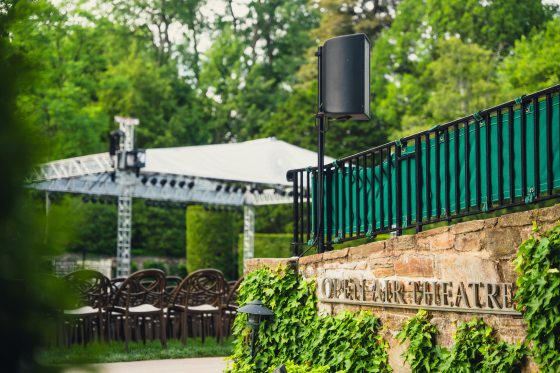 A stone half wall covered in Ivy says "Open Air Theatre." The background depicts a tree-filled setting with a white tent over a large stage with seats on a lawn.  