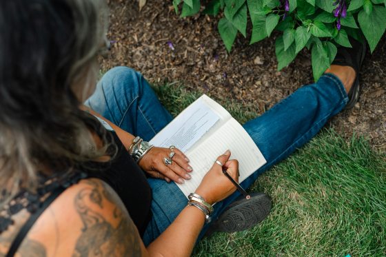 A close-up, high-angle shot showing a person sitting on the grass while writing or sketching in a notebook, with a small potted plant visible nearby.