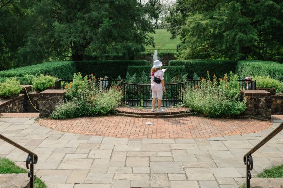 A person wearing a white hat, light pink shirt, and shorts stands on a brick patio, holding a notebook and looking towards a fountain in a lush, green garden setting.