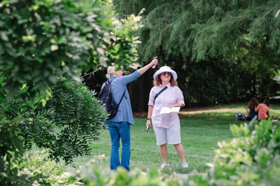 A person in a white hat and pink shirt stands in a grassy area holding a notebook, while another person beside them gestures and points upward.