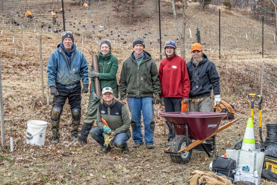 Six volunteers and staff pose for a photo on a hillside. They are dressed in heavy winter work gear, including beanies and gloves. One person holds a post driver, and a purple wheelbarrow sits to the right. The background shows a steep slope covered in protective netting and hundreds of small white tree tubes.