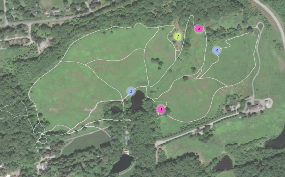 An aerial satellite view of a large green meadow and woodland area overlaid with white lines representing a network of trails. Five numbered, colorful circular markers indicate specific project locations.