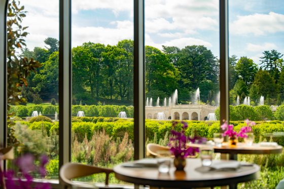 A view from a restaurant table looking out through large windows. In the background, a grand stone fountain structure with multiple water jets is surrounded by lush green trees and manicured hedges under a bright, partly cloudy sky.