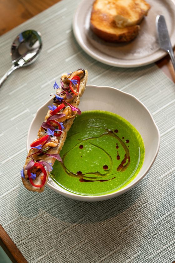 A bowl of vibrant green chilled soup topped with a swirl of dark oil. A long, thin piece of toasted bread rests across the rim, intricately garnished with mussels, sliced red chilies, purple microgreens, and small blue edible flowers.
