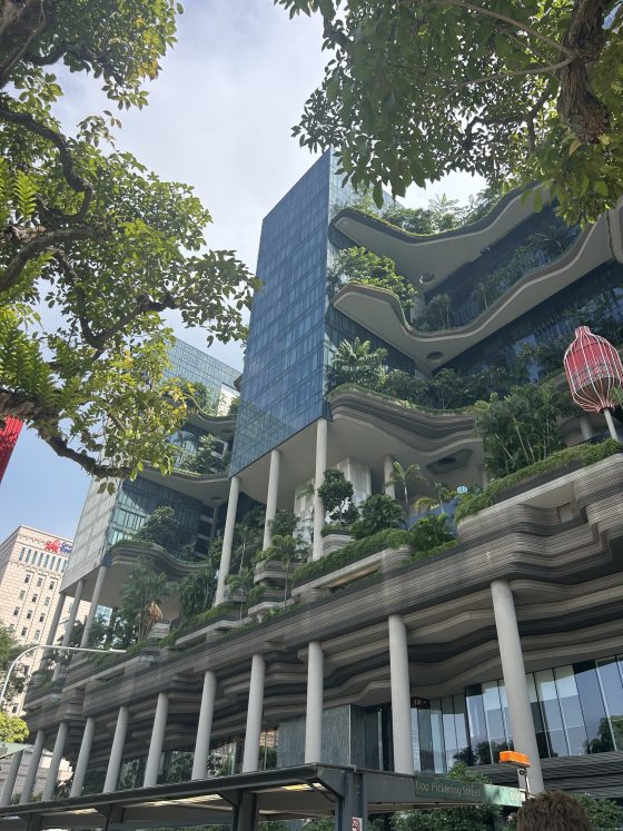 A low-angle view looking up at the unique, undulating green facade of the PARKROYAL Pickering Hotel in Singapore. The building features multiple cantilevered layers filled with tropical plants and trees, supported by tall concrete pillars against a bright sky.