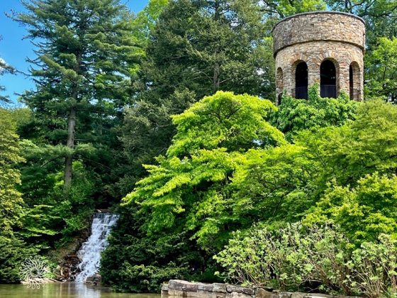 A serene garden landscape featuring a stone chimes tower and a waterfall. The tower's base is draped in climbing vines, and the surrounding foliage shows varied shades of spring green, reflecting a peaceful and immersive natural environment.
