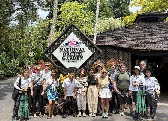 A large group photo of students and staff from the National Orchid Garden in Singapore and Longwood Gardens, posed in front of a diamond-shaped sign that reads "National Orchid Garden Singapore. They are surrounded by lush tropical greenery, vibrant orange orchids, and several individuals are holding green umbrellas.