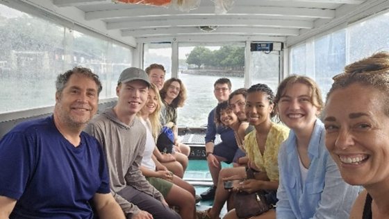 A candid group photo taken from inside a small, white-roofed ferry boat moving across the water. On the far left, Chad Davis (Associate Vice President of Conservatories) smiles at the camera, wearing a blue t-shirt. On the far right, Lauren Hill (Domestic & International Studies Manager) beams in a close-up selfie-style angle. Between them, two rows of students from the Professional Horticulture class of 2026 sit facing each other, all smiling as they travel toward the lush, green shoreline of Pulau Ubin vi