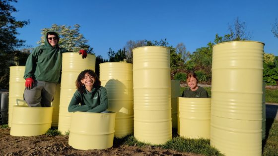 Three people posing alongside yellow barrels in varying heights.