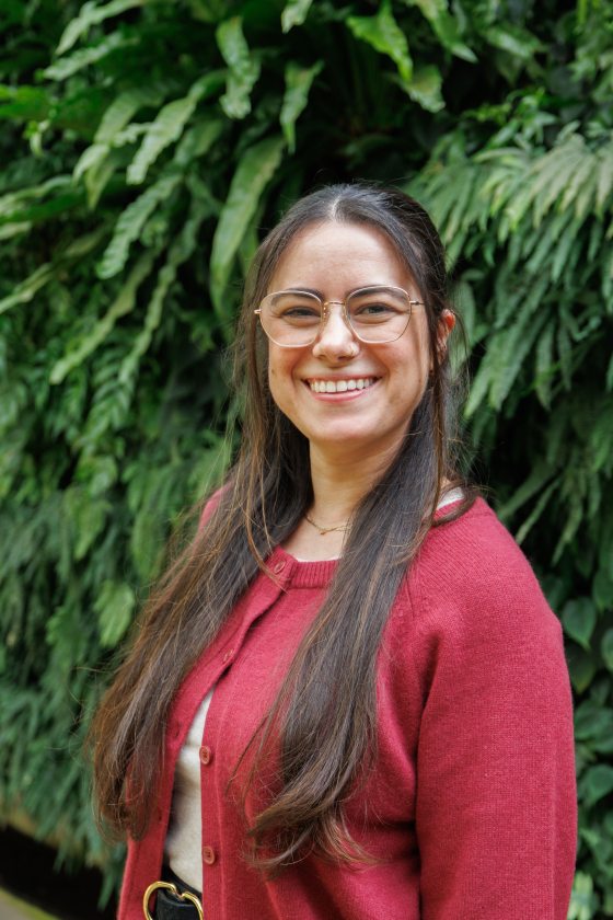 A woman with dark hair and glasses in a burgundy cardigan smiles in front of a lush green wall. 