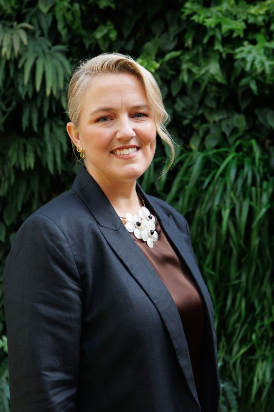 A woman with blonde hair wearing a navy blazer, brown satin shirt, and white flower necklace smiles in front of a lush green wall. 
