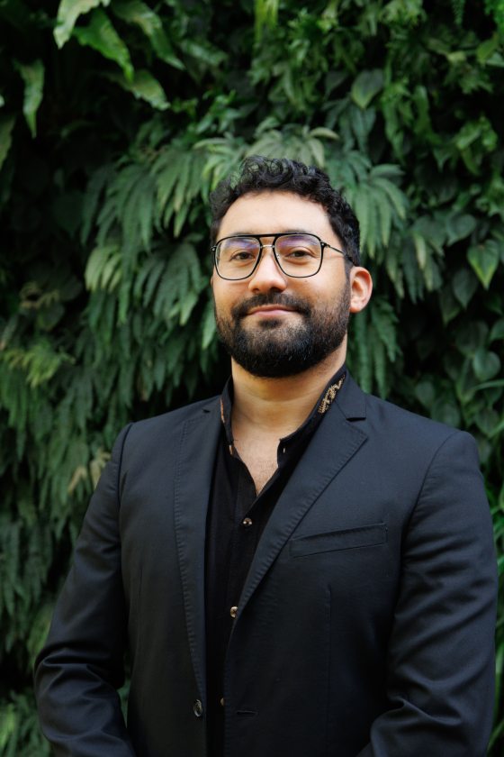 A man with black hair, a short beard, and black glasses in a black suit poses in front of a lush green wall.  