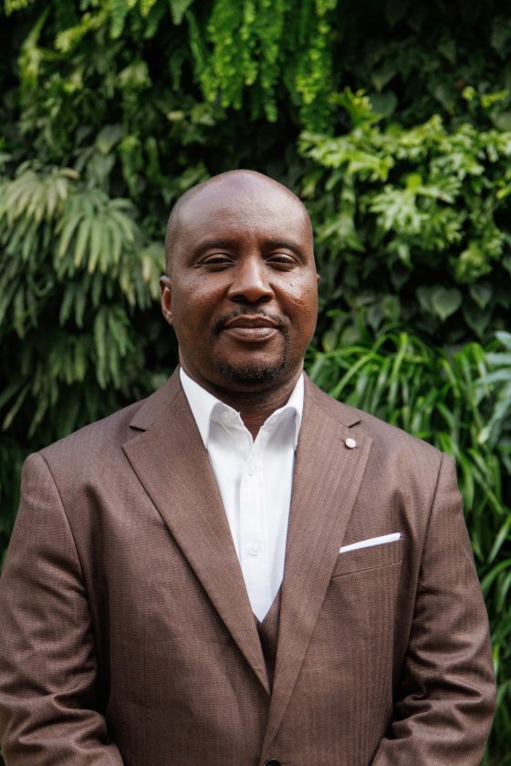 A man wearing a brown suit with white shirt poses in front of a lush green wall 