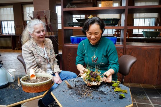 Two people sitting together at a workshop table covered in loose soil, actively working on a plant arrangement. The woman on the right, in a teal sweater, is meticulously planting moss into a shell-like Kusamono pot. The woman on the left, in a patterned blazer, watches with a smile. Nearby are a container of soil mix, small scissors, a brush, and patches of fresh green moss waiting to be used.
