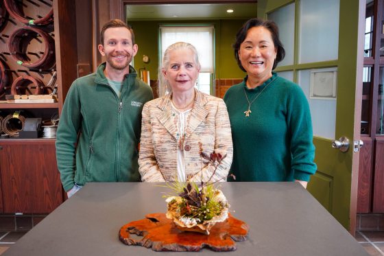 A group portrait of three smiling people standing side-by-side behind a gray table. On the table sits a completed Kusamono arrangement in a light-colored, ruffled ceramic shell-like pot placed on a polished, natural live-edge wood slab. The person on the left wears a green fleece jacket with a "Longwood Gardens" logo, the person in the center wears a patterned tan blazer, and the person on the right wears a dark teal sweater.