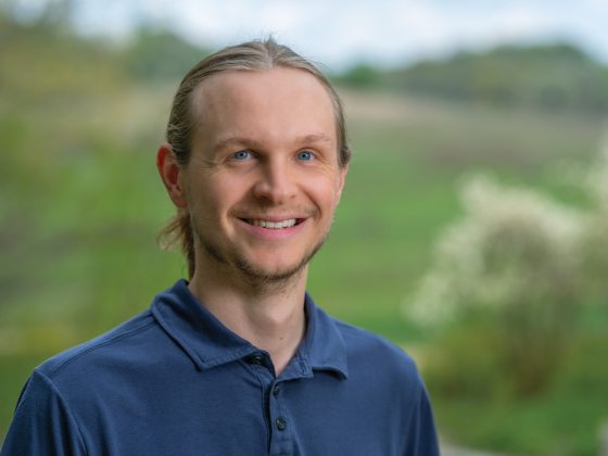 A man with blonde hair wearing a navy shirt smiles in front of a green and grassy meadow. 