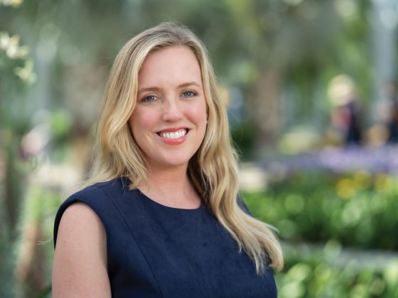 A woman with blonde hair wearing a navy shirt smiles in front of a backdrop of lush green plants 
