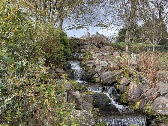 A scenic, multi-tiered rock waterfall cascading down a lush hillside. The stream is surrounded by dense green shrubs, budding trees, and rocky terrain. The rooftop of a traditional building is visible peeking over the top of the hill against an overcast sky.