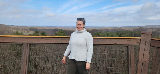 A woman smiling while standing on an elevated wooden canopy walk or observation deck. She is wearing a cozy grey turtleneck sweater and dark pants. The background features an expansive, rolling view of a dense forest stretching to the horizon under a cloudy sky.
